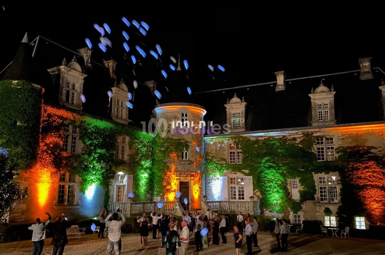 Des personnes libèrent des ballons lumineux devant un château éclairé de nuit par des projecteurs colorés.