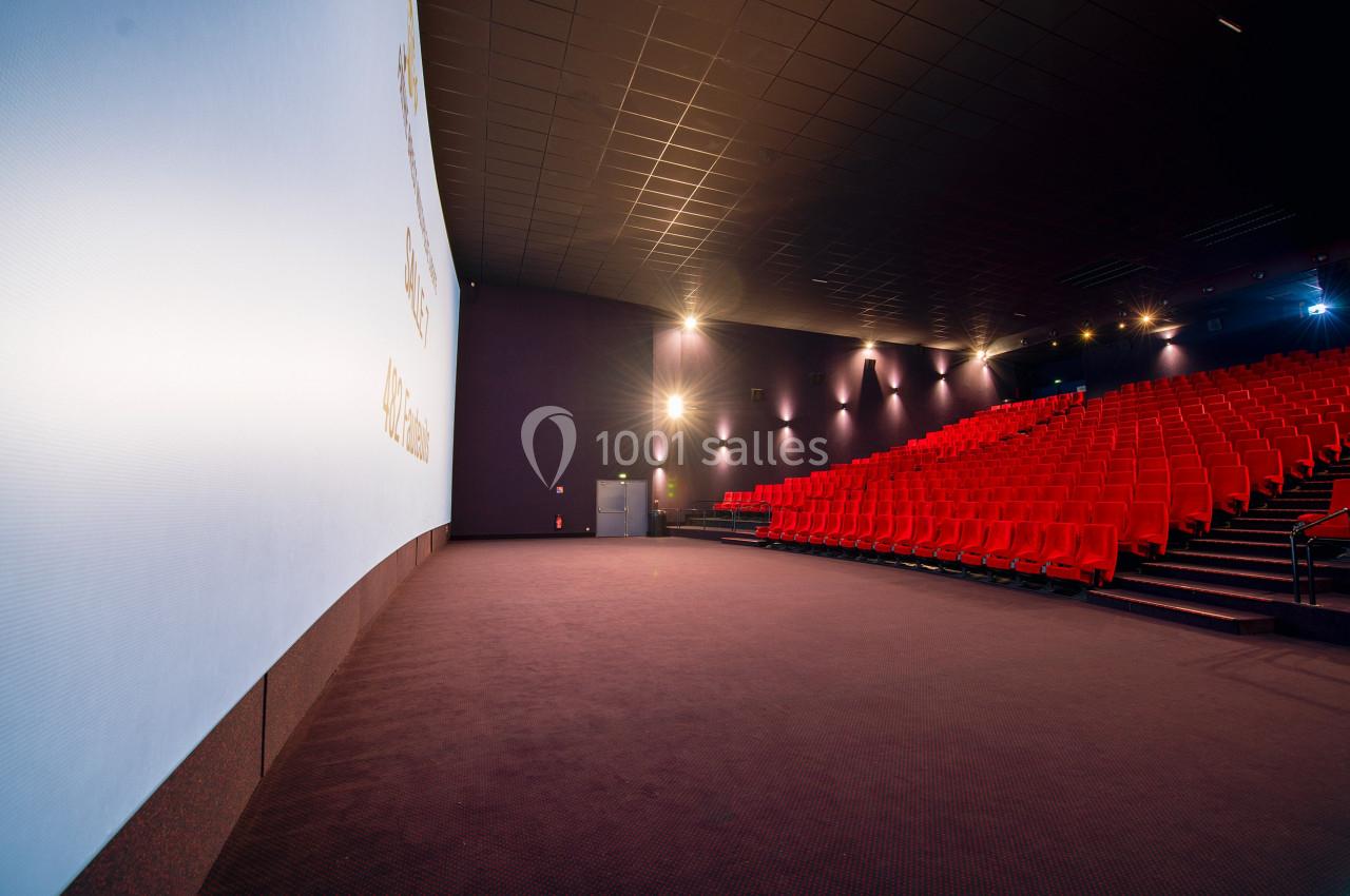 Salle de cinéma vide avec écran large, rangées de sièges rouges et éclairage tamisé.