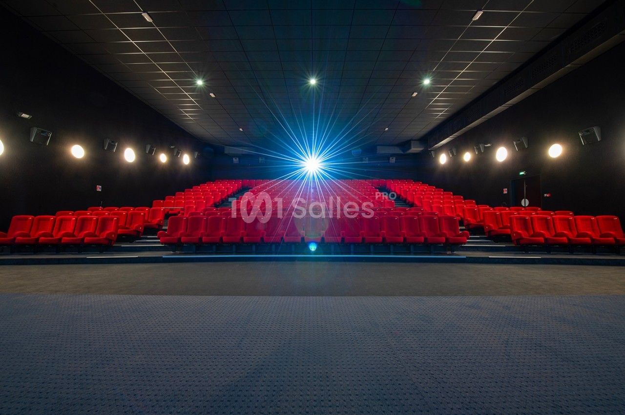 Salle de cinéma vide avec des rangées de sièges rouges et un projecteur allumé au fond.
