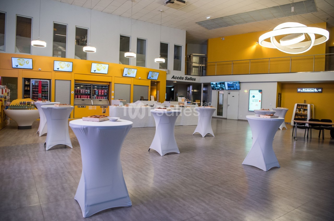 Salle lumineuse avec tables hautes habillées de nappes blanches, décorée de murs jaunes et écrans affichant des images.