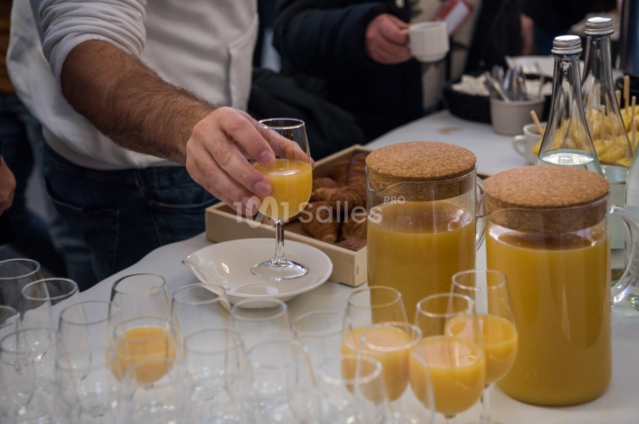Une personne sert un verre de jus d'orange sur une table garnie de carafes, verres et viennoiseries.