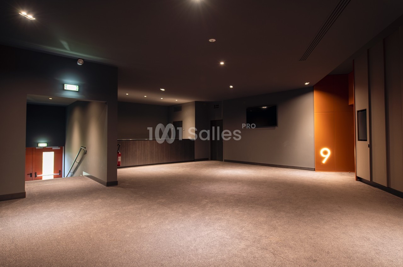 Hall d'entrée d'un cinéma avec moquette, éclairage tamisé, écran mural et numéro 9 lumineux sur un mur orange.