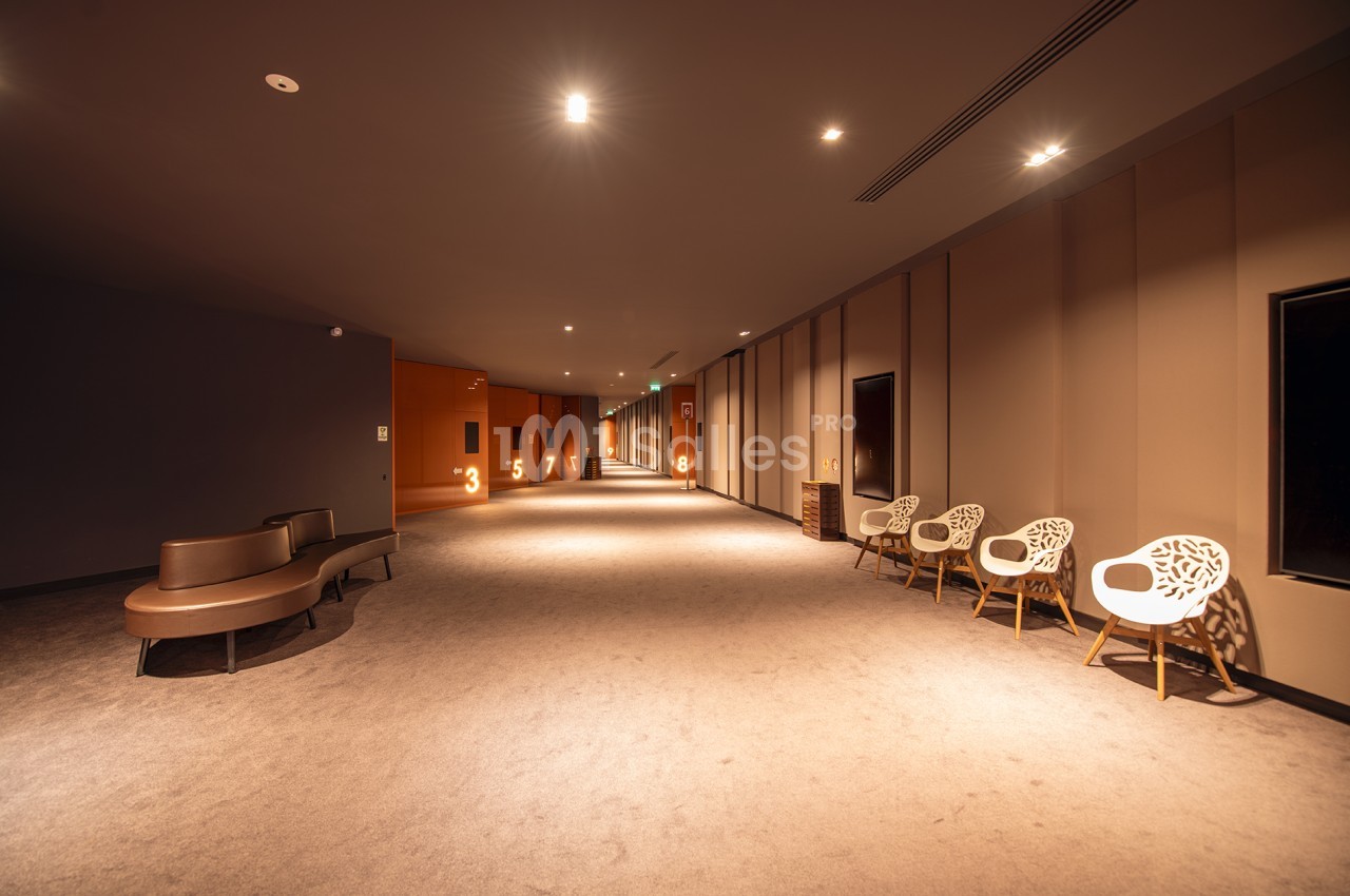 Couloir moderne avec moquette, éclairage tamisé, sièges design et numéros de salles visibles sur les portes.