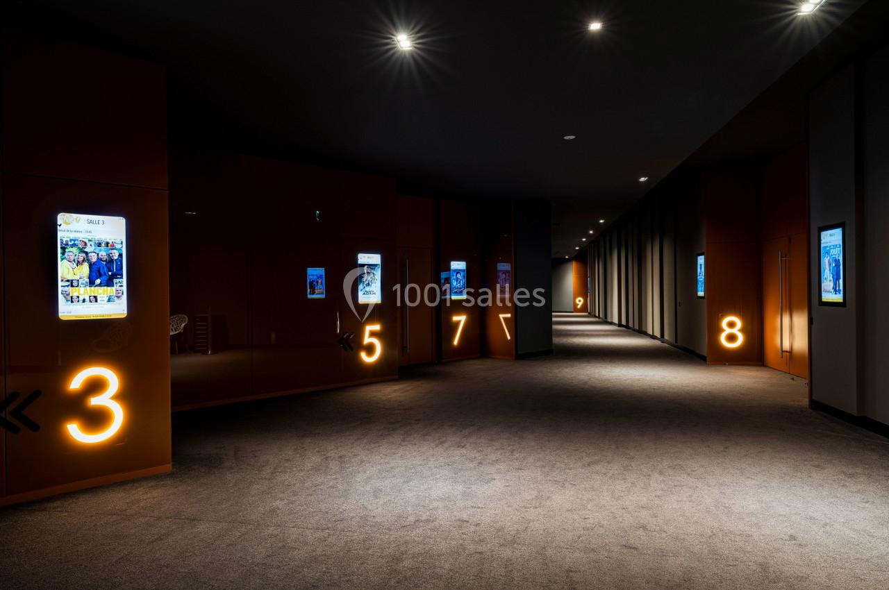 Couloir sombre avec moquette, numéros lumineux sur les murs et affiches rétroéclairées alignées.