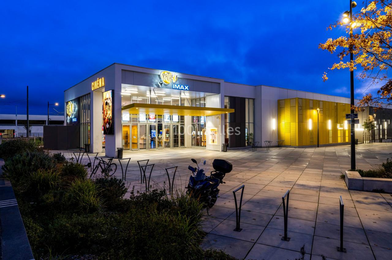 Façade d'un cinéma moderne avec enseigne IMAX, éclairée en soirée, entourée de végétation et d'un parking.