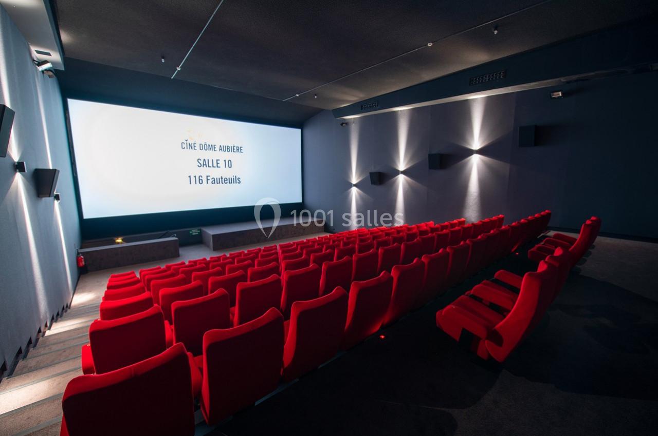 Salle de cinéma moderne avec rangées de fauteuils rouges, écran blanc affichant des informations sur la salle.