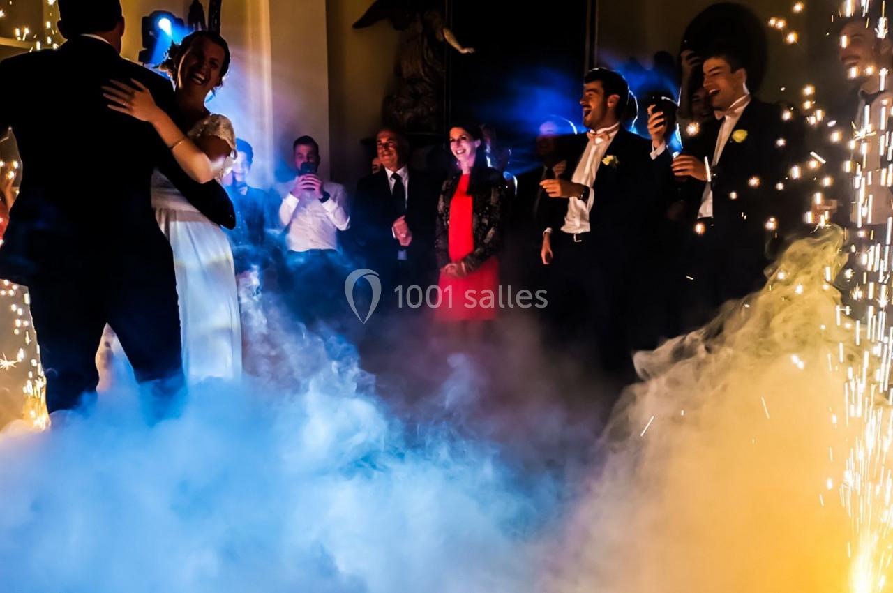 Un couple danse au milieu de la fumée et des étincelles, entouré d'invités élégants dans une ambiance festive.