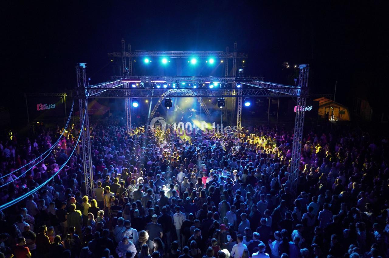 Foule rassemblée devant une scène illuminée par des projecteurs bleus lors d'un concert en plein air de nuit.