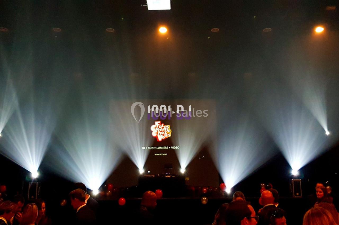 Salle sombre avec scène éclairée par des projecteurs, écran affichant un logo coloré et des personnes en contrebas.