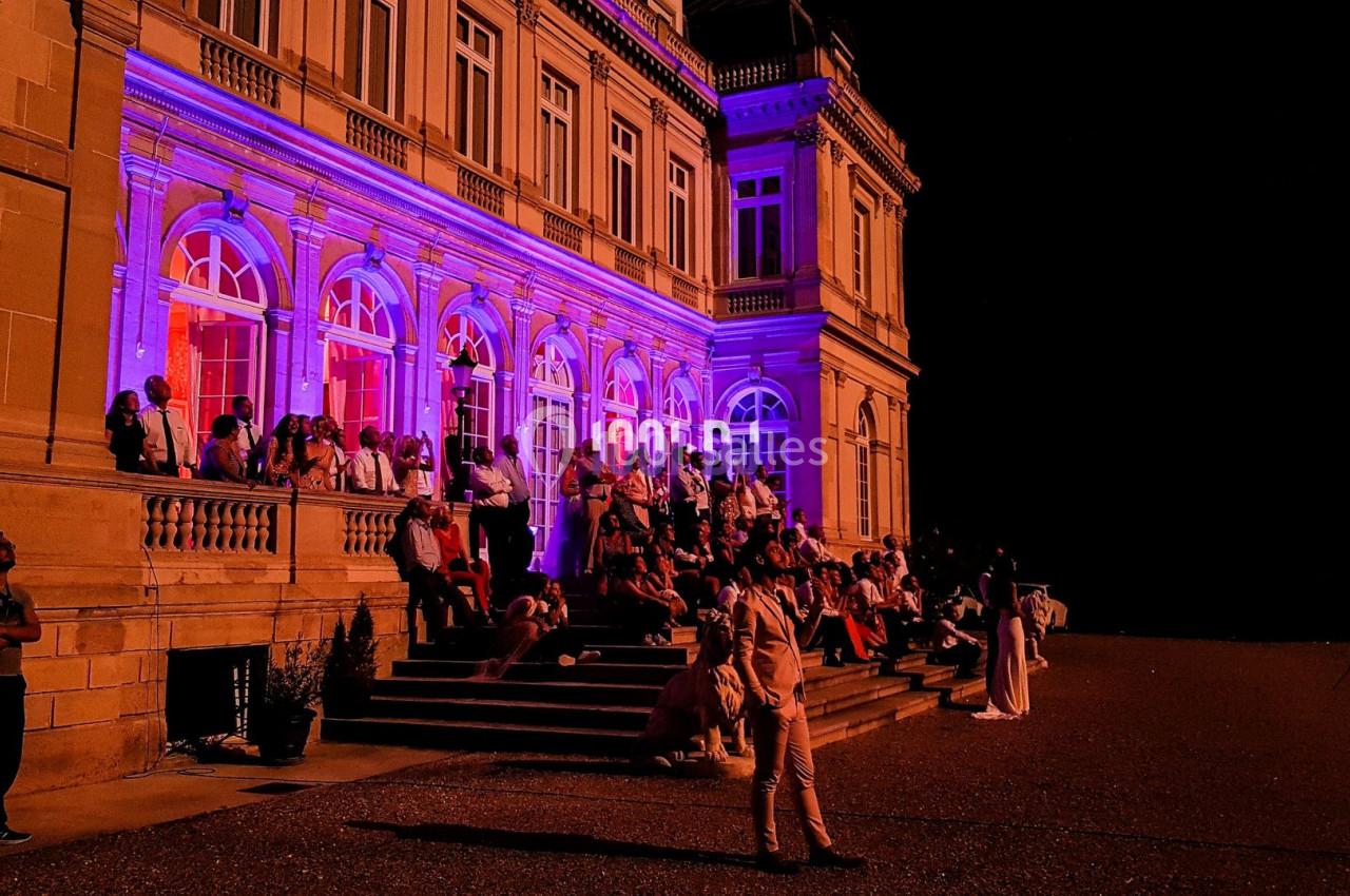 Façade d'un bâtiment illuminé en violet avec des personnes assises et debout sur les marches, de nuit.