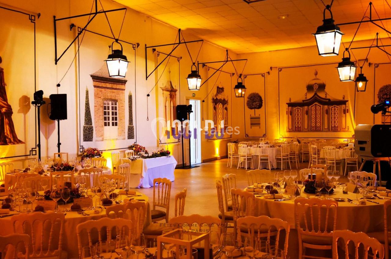Salle de réception décorée avec des tables rondes dressées, des chaises en bois clair et des éclairages suspendus.
