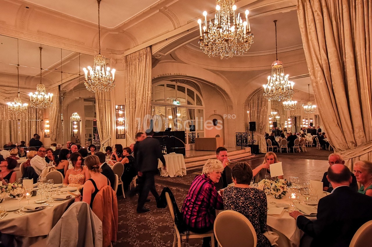 Salle de réception élégante avec des invités dînant à des tables rondes, entourés de lustres et de rideaux luxueux.