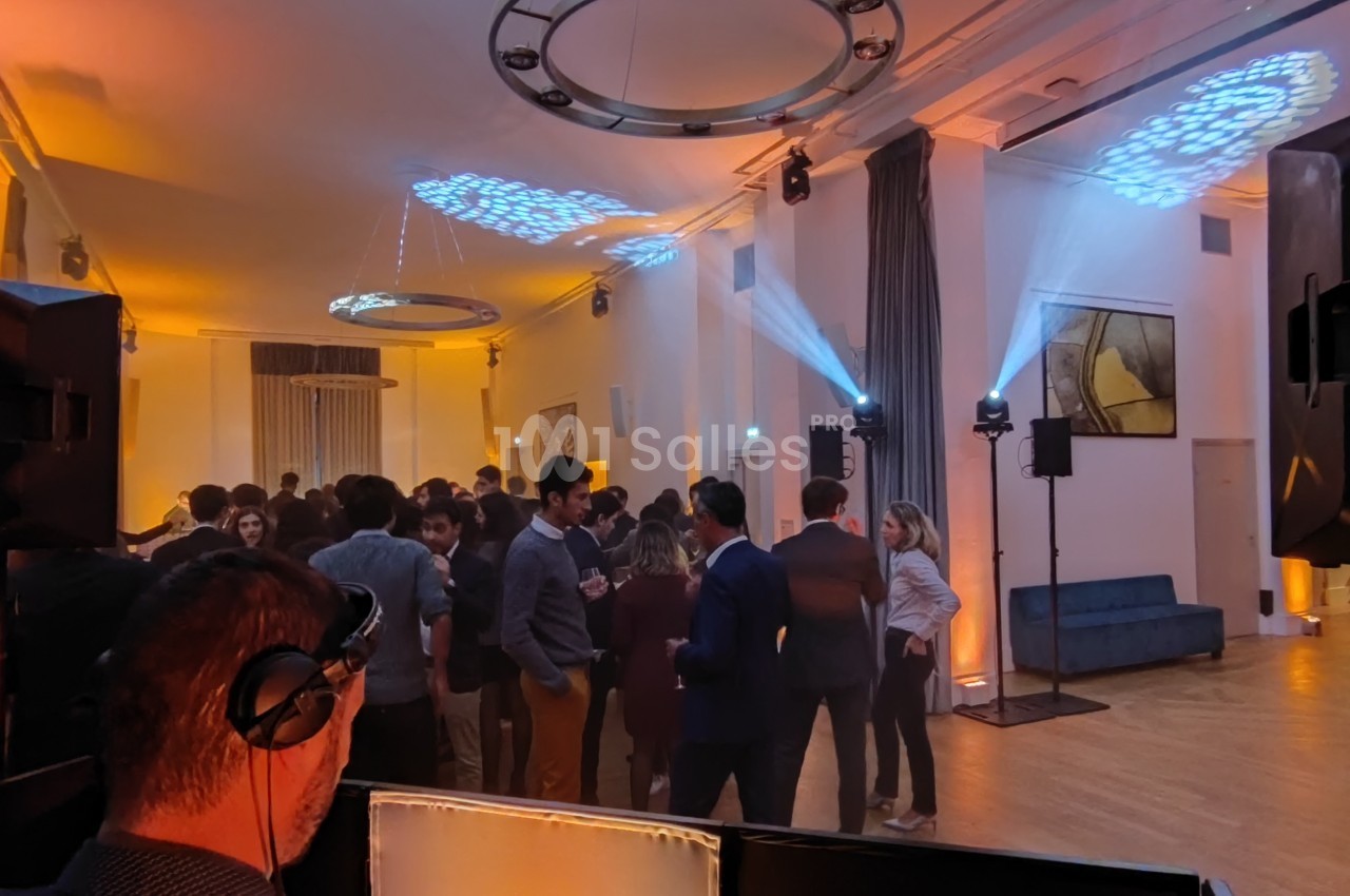 Salle de réception avec un DJ au premier plan, des invités discutant et des jeux de lumière colorés au plafond.