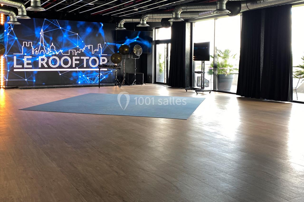 Salle lumineuse avec parquet, écran géant affichant ’Le Rooftop’, projecteurs et grandes baies vitrées donnant sur une…