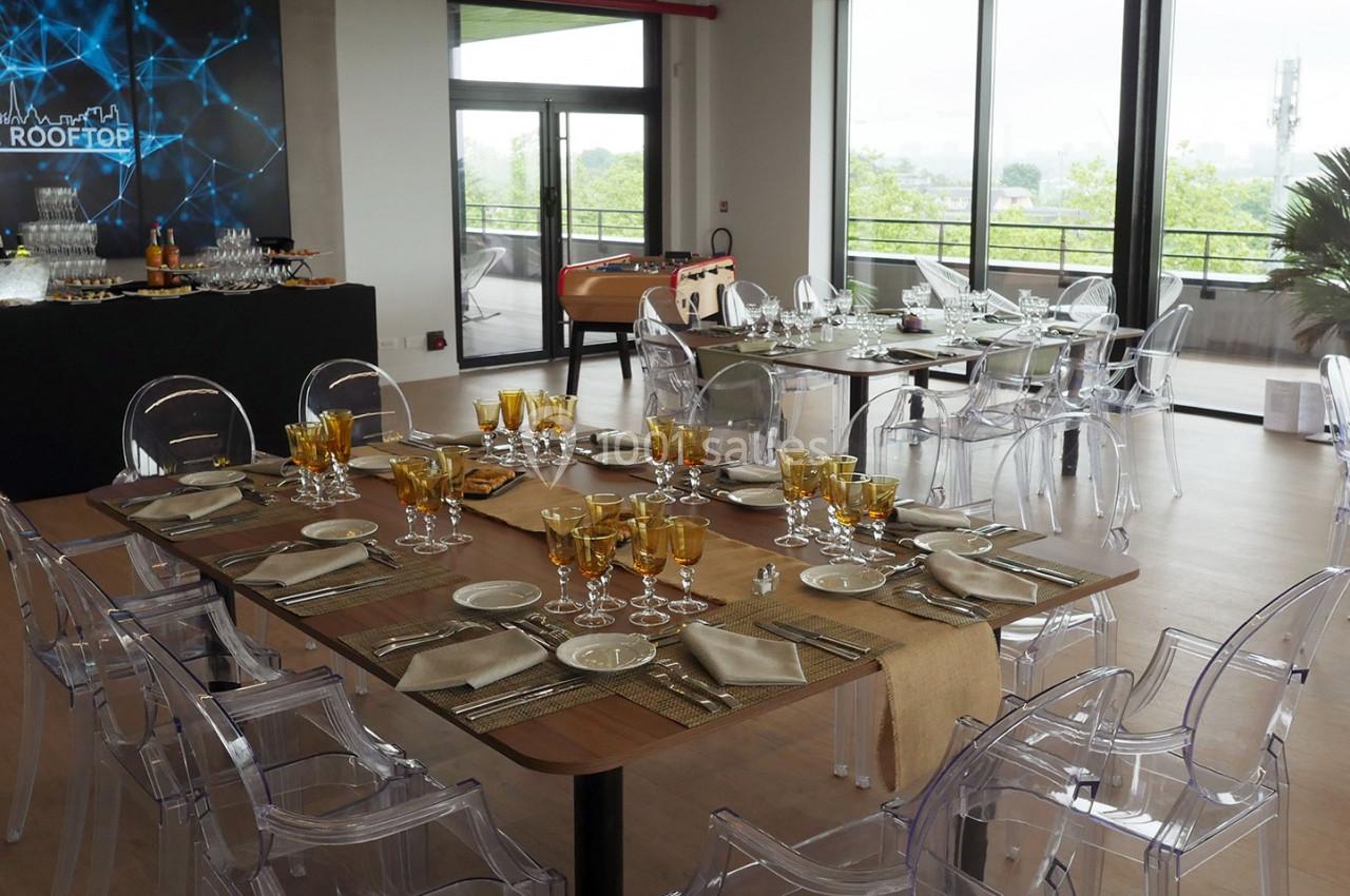 Salle lumineuse aménagée pour un événement, avec tables dressées, chaises transparentes et vue sur une terrasse extérieure.