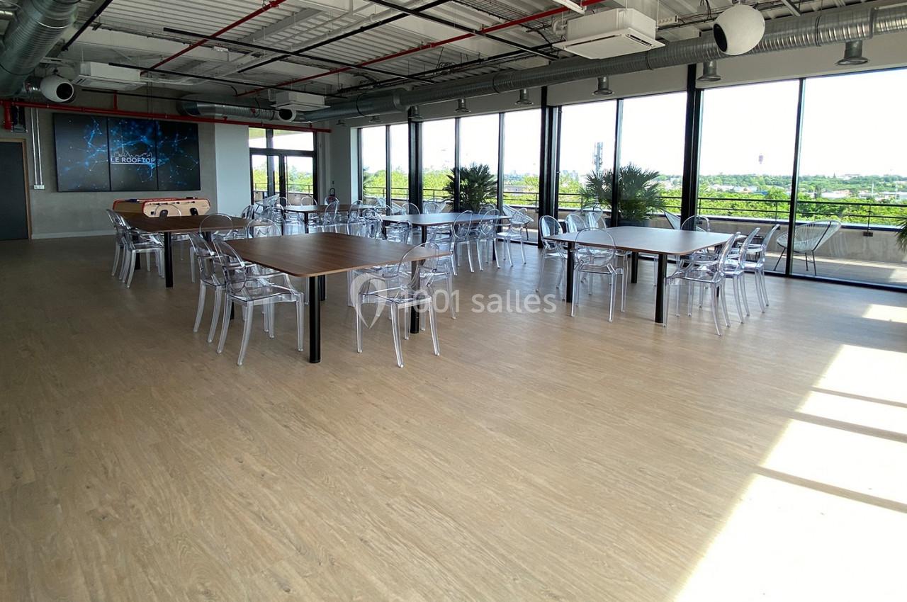 Salle lumineuse avec grandes baies vitrées, tables en bois, chaises transparentes et vue sur un espace extérieur verdoyant.