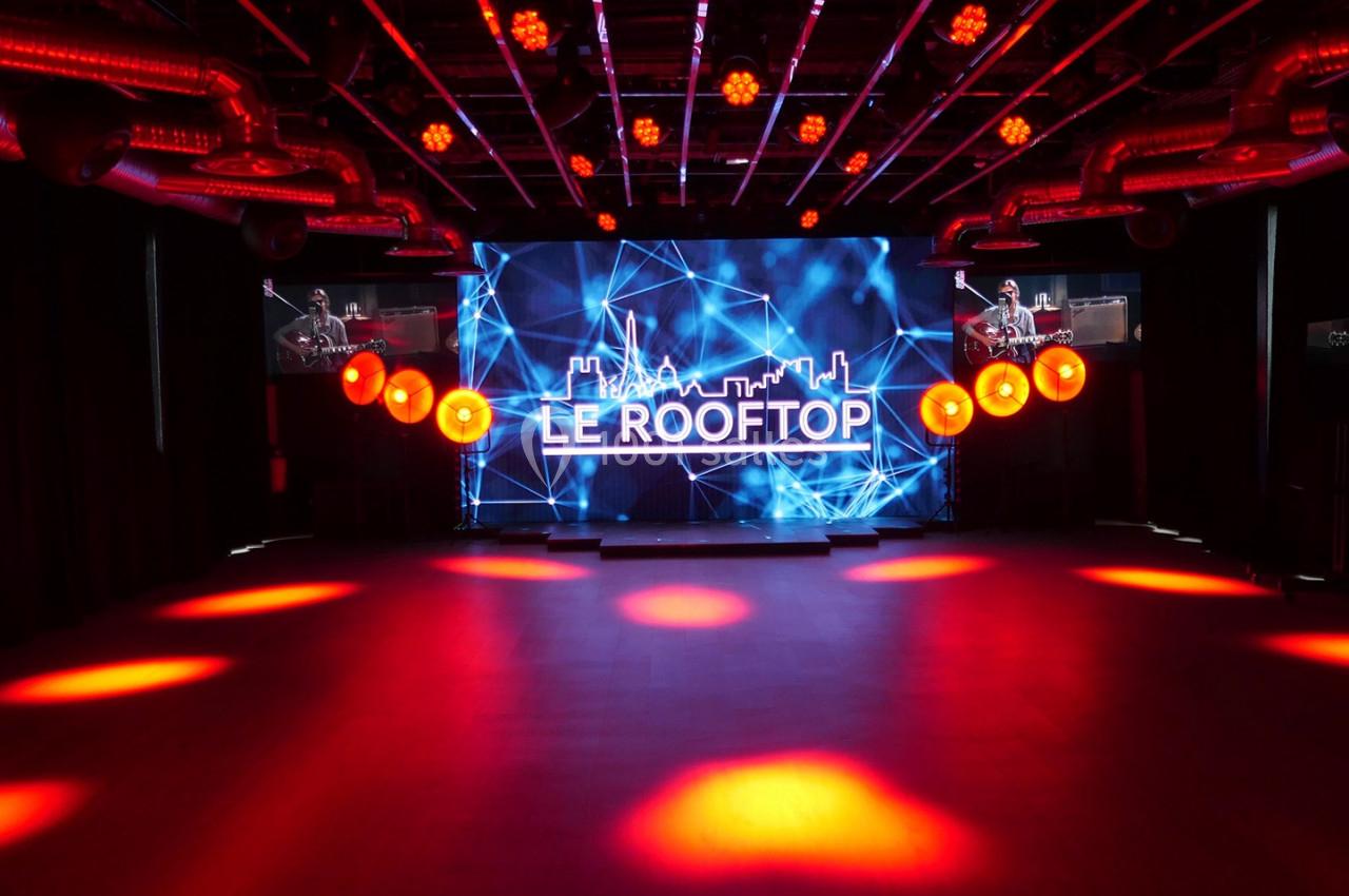 Salle sombre avec éclairage rouge, grand écran affichant ’Le Rooftop’ et décorations lumineuses suspendues.