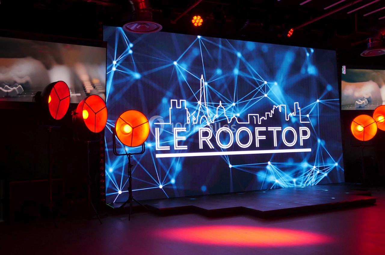 Écran lumineux affichant ’Le Rooftop’ entouré de projecteurs orange dans un espace intérieur sombre.