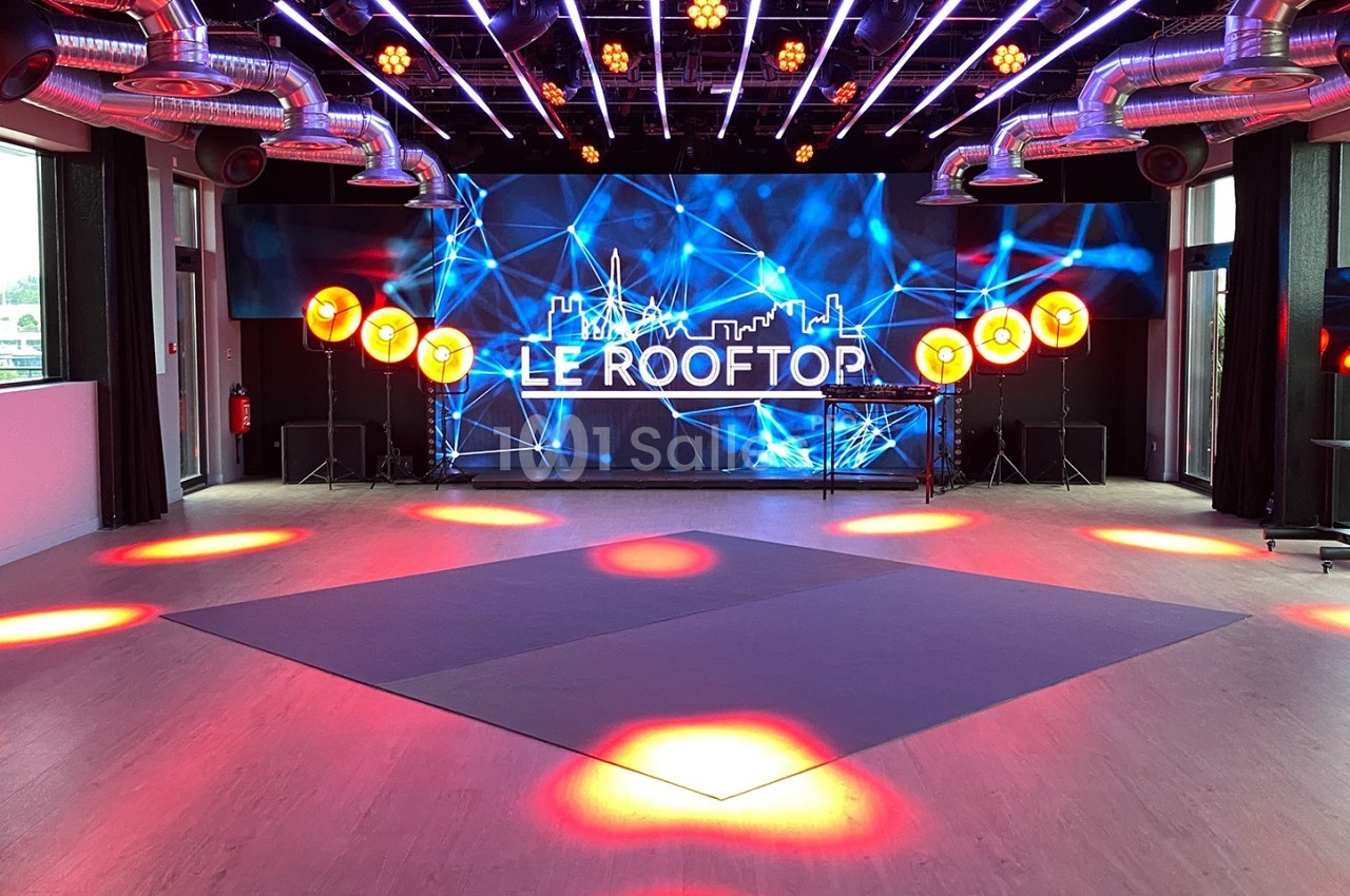 Salle lumineuse avec écran géant affichant ’Le Rooftop’, éclairages colorés et grandes baies vitrées.