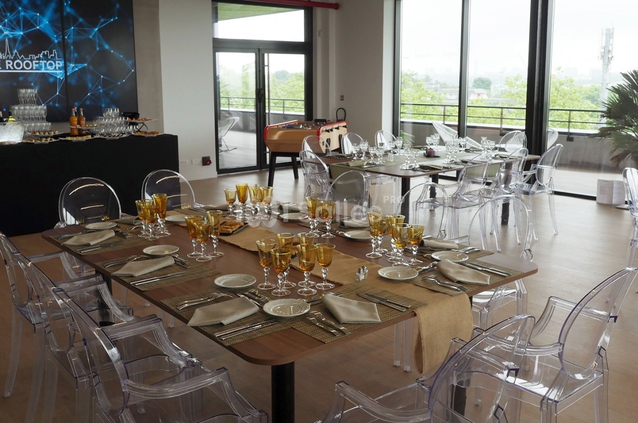 Salle lumineuse aménagée pour un événement, avec tables dressées, chaises transparentes et vue sur une terrasse arborée.