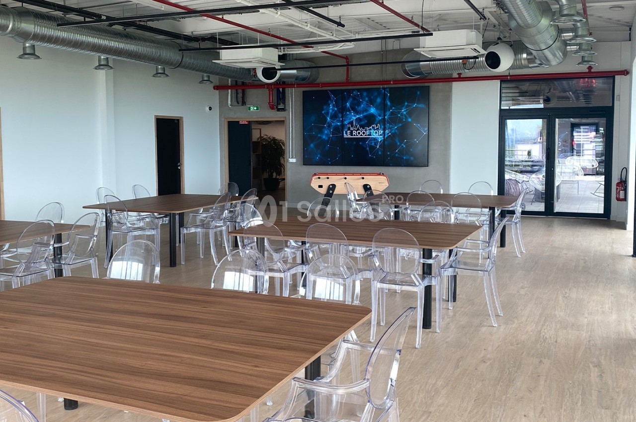 Salle lumineuse avec tables en bois, chaises transparentes, écran mural, baby-foot et grandes baies vitrées.