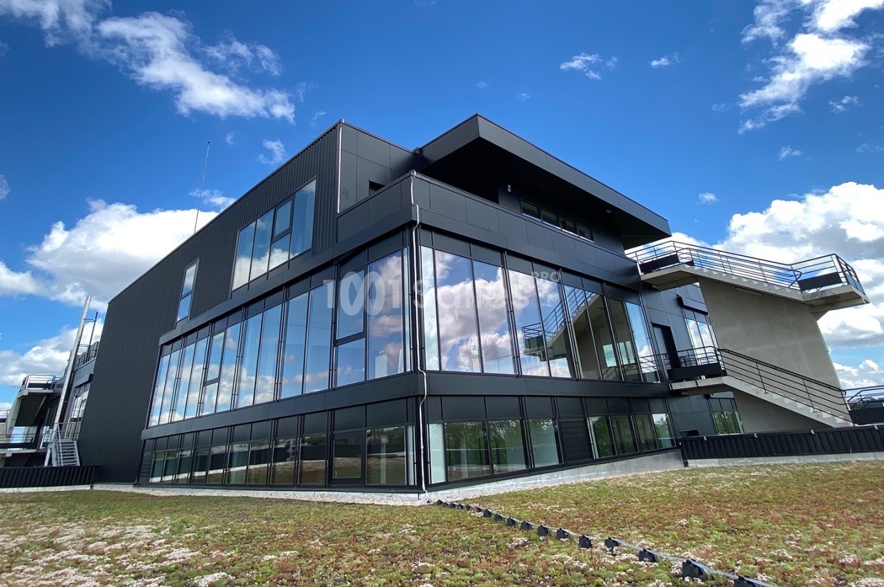 Bâtiment moderne à façade vitrée, entouré de végétation sur un toit, sous un ciel bleu avec quelques nuages.