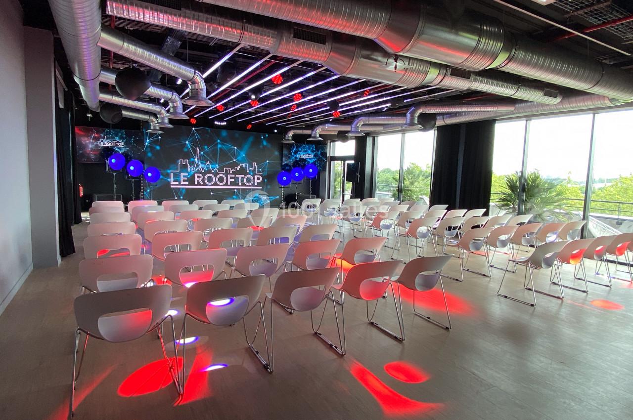 Salle de conférence moderne avec rangées de chaises blanches, éclairage coloré et grande baie vitrée donnant sur l'extérieur.