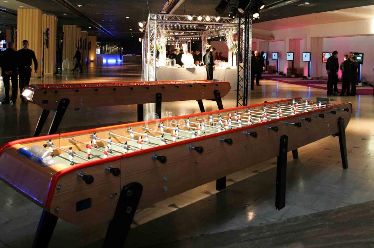 Deux baby-foot en bois éclairés dans un espace intérieur moderne avec des invités et des stands en arrière-plan.