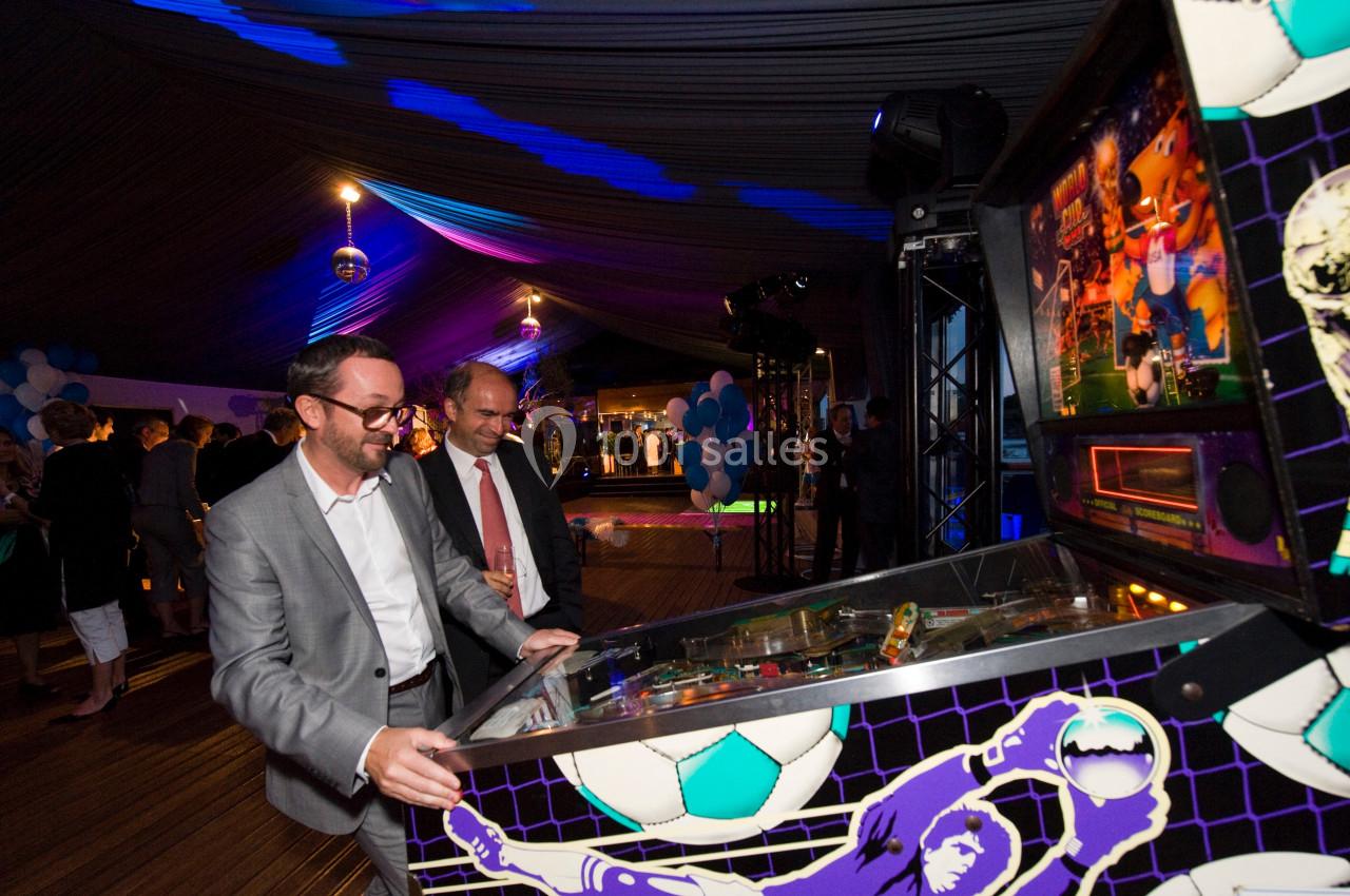 Deux hommes en costume jouent à un flipper sur le thème du football dans une salle éclairée de lumières colorées.