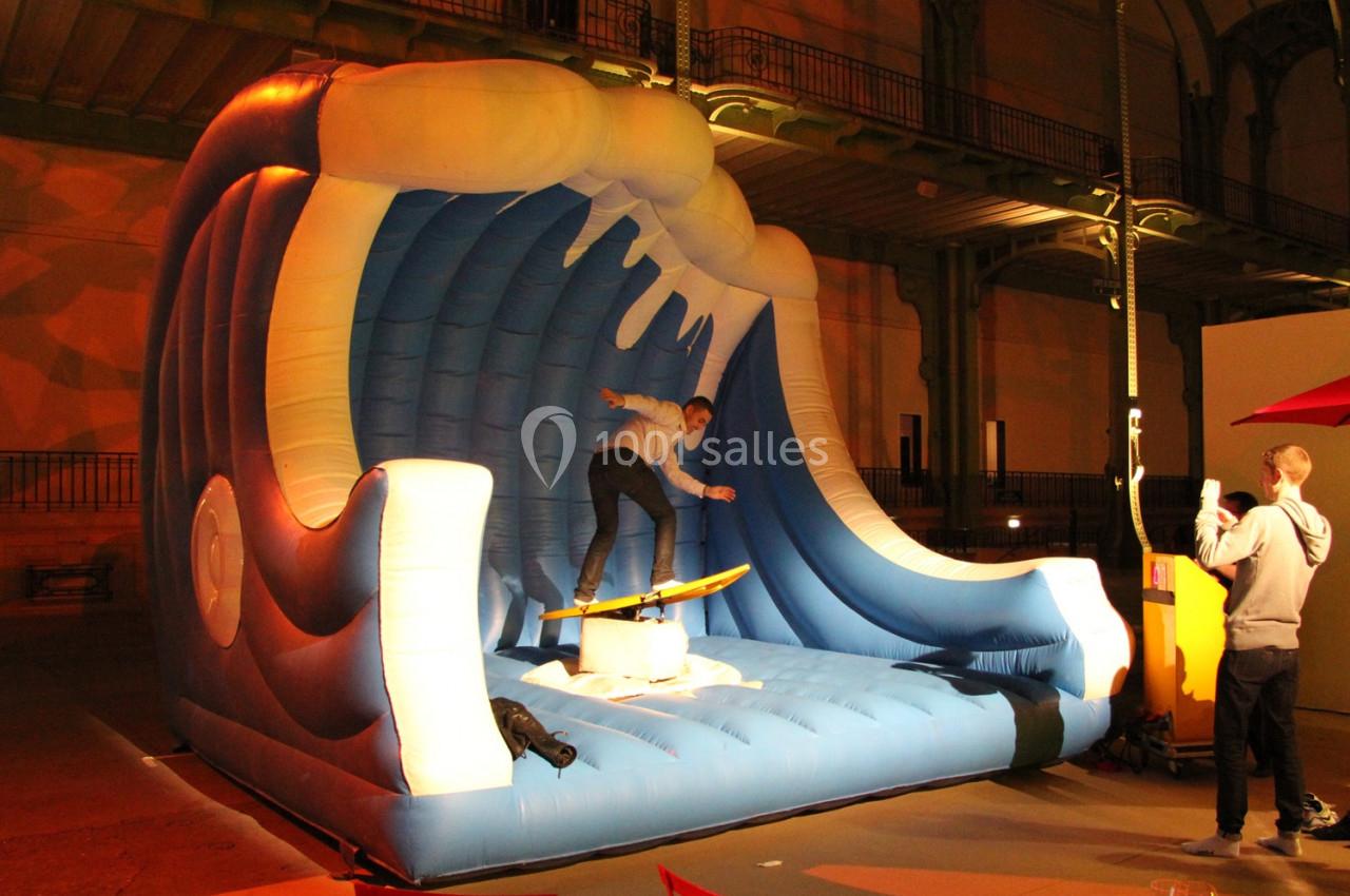 Homme sur une planche de surf simulant une vague géante gonflable dans un espace intérieur éclairé.