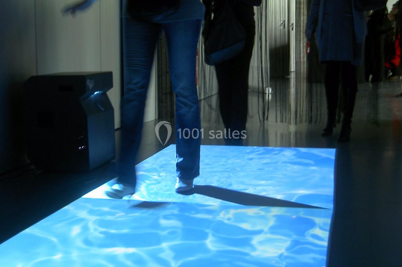 Personne marchant sur une projection lumineuse simulant une surface d'eau, dans un espace intérieur sombre.