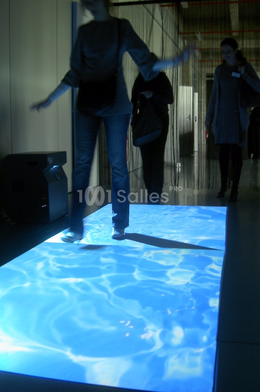 Personne marchant sur une projection lumineuse simulant une surface d'eau, dans un espace intérieur sombre.
