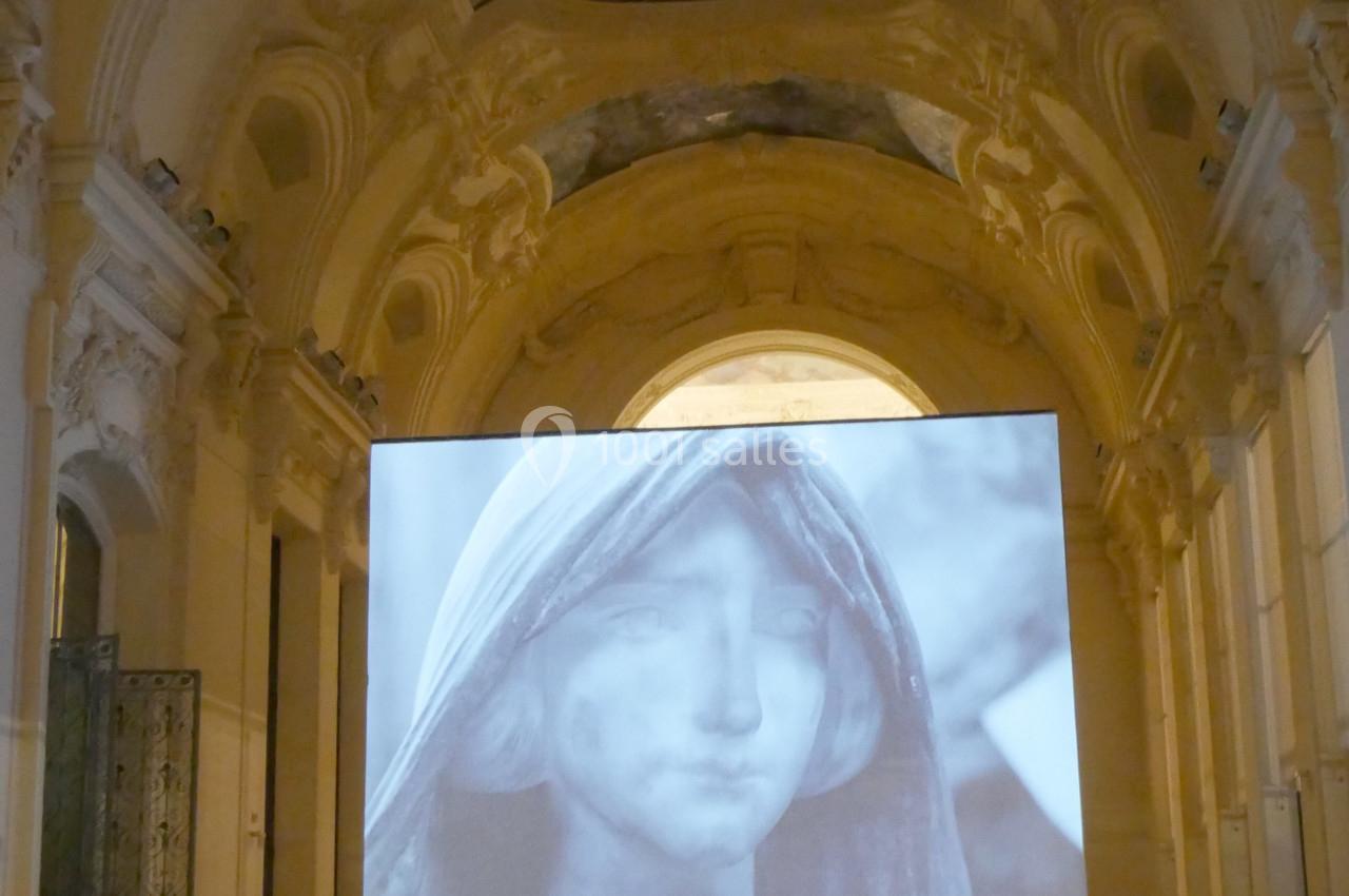 Projection d'une image en noir et blanc d'un visage voilé dans une grande salle ornée d'architecture classique.