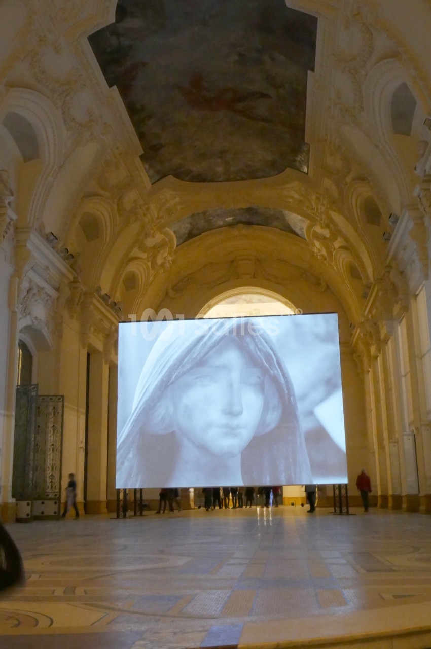 Projection d'une image en noir et blanc d'un visage voilé dans une grande salle ornée d'architecture classique.