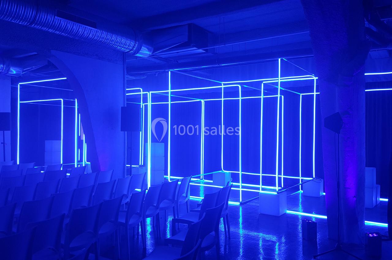 Installation lumineuse composée de structures géométriques en néons bleus dans une salle avec des sièges alignés.