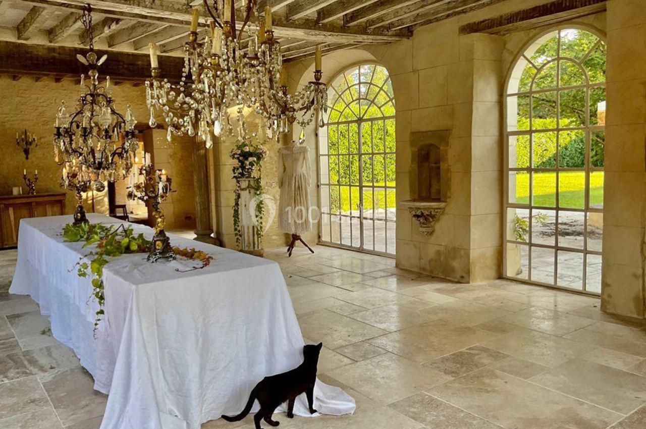 Salle lumineuse avec grandes fenêtres, table blanche décorée, lustres suspendus et un chat noir au sol.
