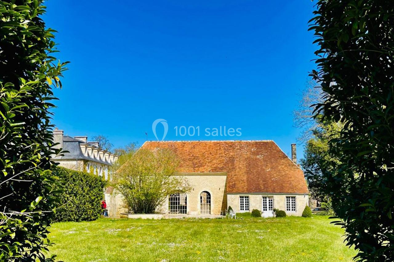 Vue d'une maison en pierre avec un toit en tuiles rouges, entourée de verdure sous un ciel bleu dégagé.