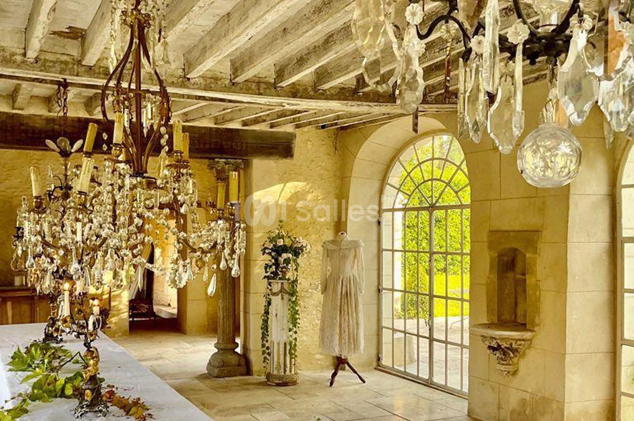 Salle lumineuse avec murs en pierre, poutres apparentes, lustres en cristal, table décorée et robe sur un mannequin.