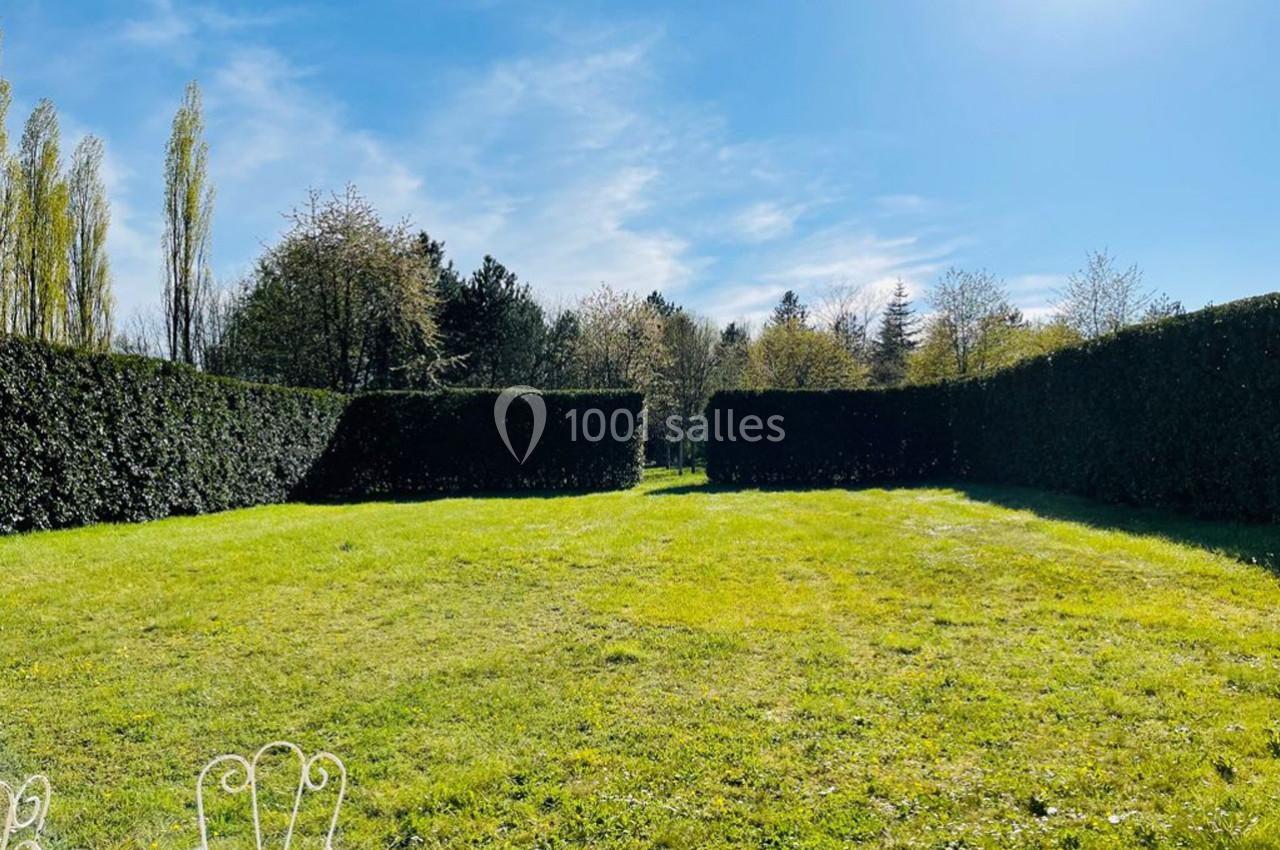 Pelouse verte entourée de haies taillées, avec des arbres en arrière-plan sous un ciel bleu ensoleillé.