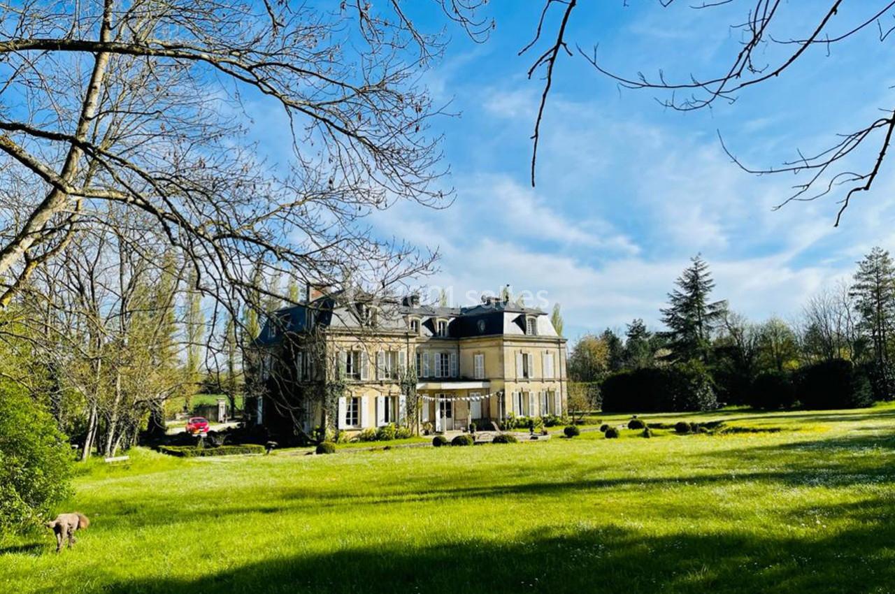 Manoir entouré d'un grand jardin verdoyant avec des arbres, sous un ciel bleu clair.