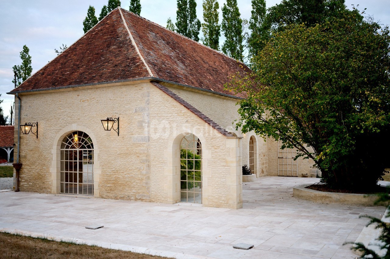 Bâtiment en pierre avec toit en tuiles rouges, grandes fenêtres en arc et terrasse pavée entourée de verdure.
