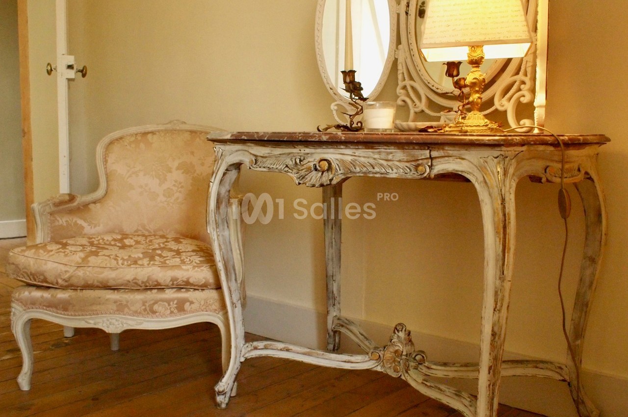 Fauteuil et console en bois sculpté de style ancien, avec une lampe allumée et un miroir, dans une pièce au sol en bois.