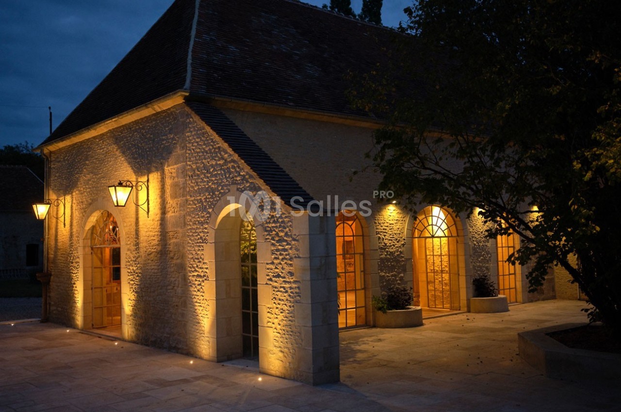 Bâtiment en pierre éclairé par des lampes extérieures, avec des arches vitrées, dans un cadre nocturne.
