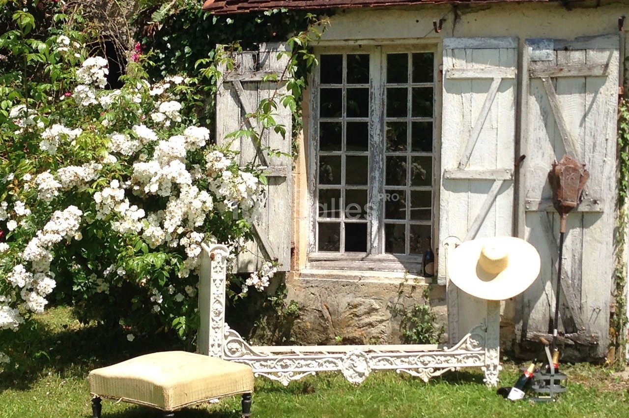 Petite maison en pierre avec volets en bois, entourée de fleurs blanches, un banc et un chapeau posés devant.