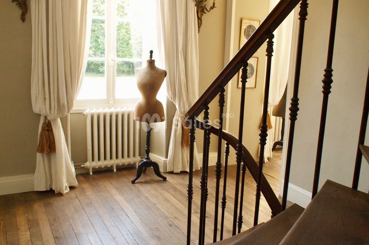 Escalier en bois menant à une pièce lumineuse avec un mannequin de couture près d'une fenêtre encadrée de rideaux blancs.