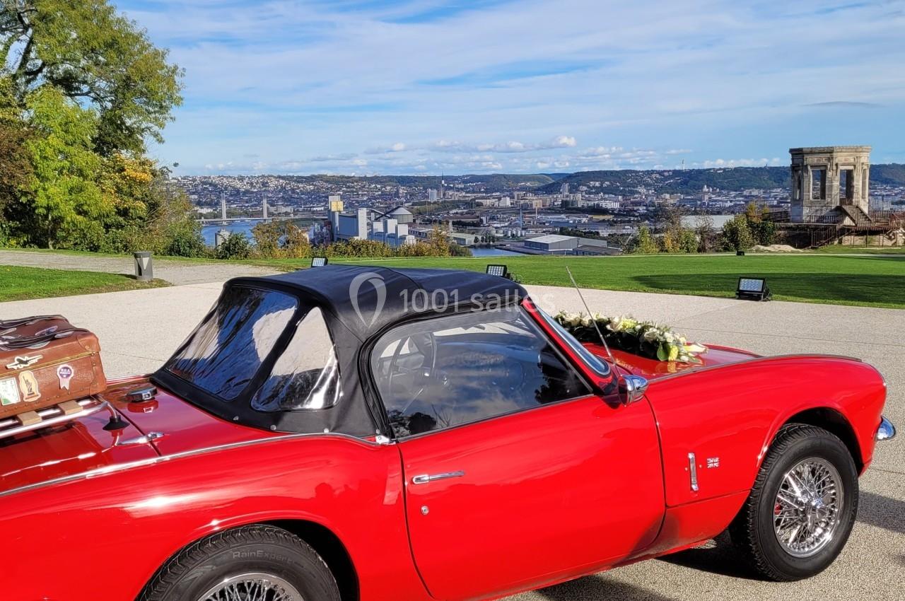 Château des Deux Lions - Exposition Voiture décapotable rouge avec valise ancienne sur le coffre, stationnée devant un panorama urbain et un monument.