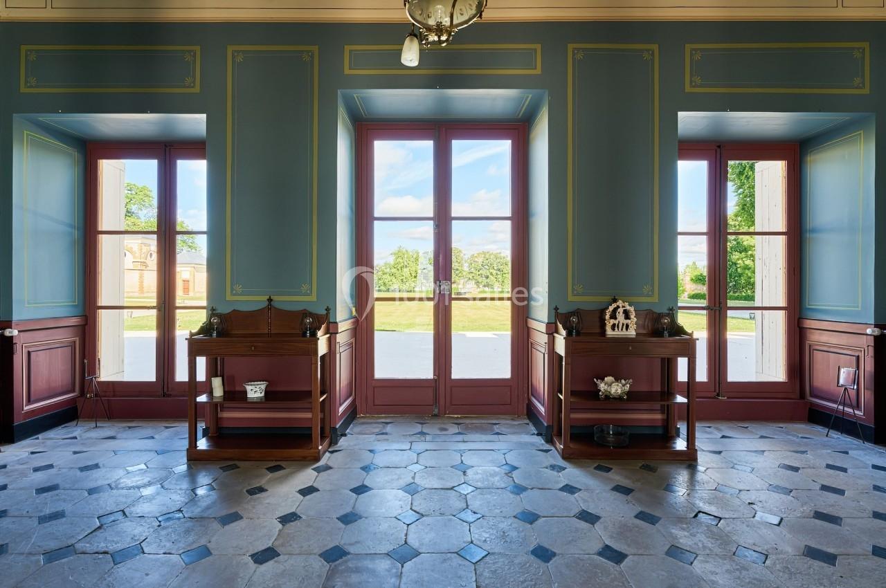 Château des Deux Lions - Hall RDC accueil Salle lumineuse avec carrelage ancien, murs bleus ornés de moulures, et grandes portes vitrées donnant sur un jardin.