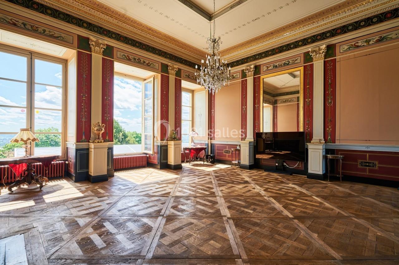 Château des Deux Lions - Salon magistral (partie restauration ou réunion avec écran) Salle élégante avec parquet en bois, murs ornés de moulures rouges et dorées, lustre central et grandes fenêtres lumineuses.