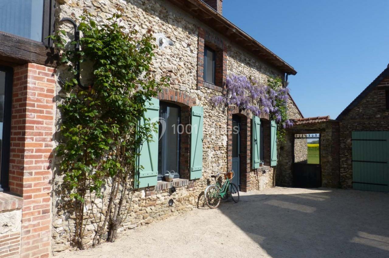 Façade en pierre d'une maison avec volets verts, glycine en fleurs et une bicyclette posée devant.