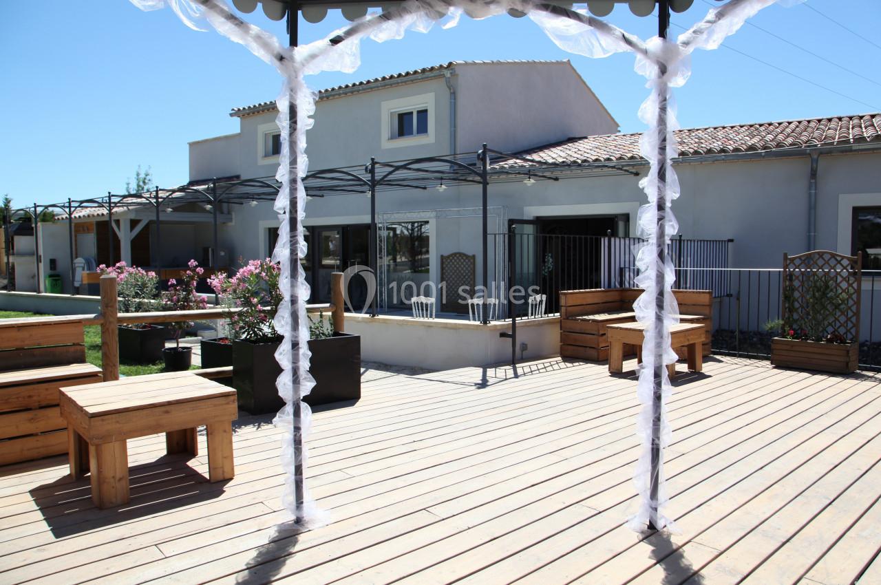 Terrasse en bois avec pergola décorée, tables et chaises, devant une maison moderne sous un ciel dégagé.